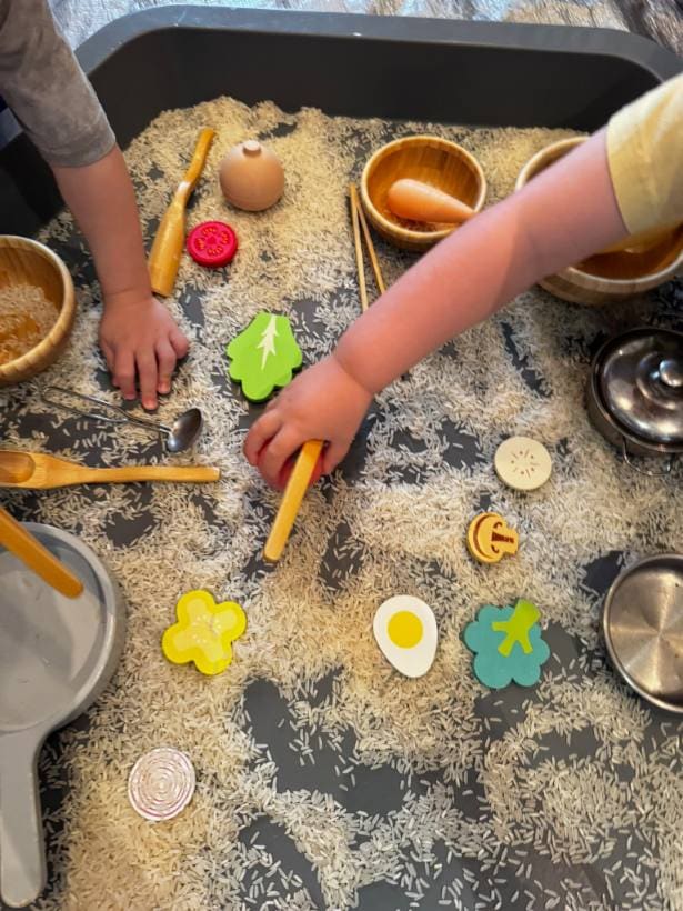 Mãos de criança explorando uma bandeja sensorial com arroz e utensílios